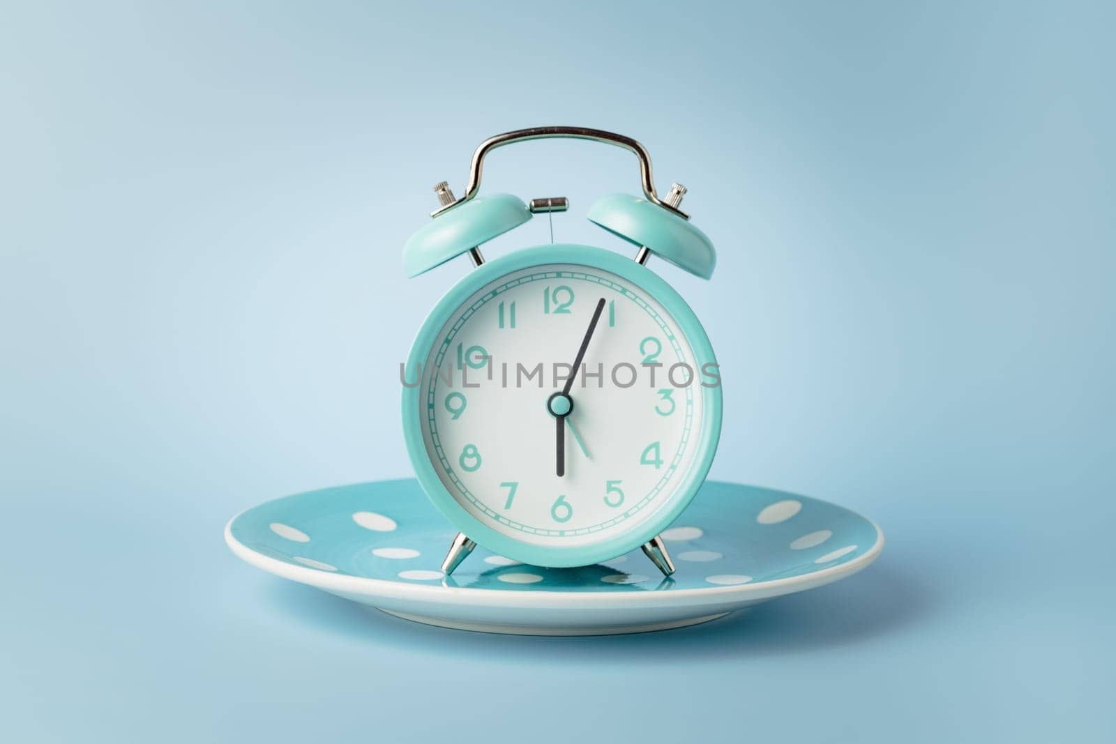 An alarm clock on an empty plate against blue background for the concept of food, time management, losing weight and eating on time.