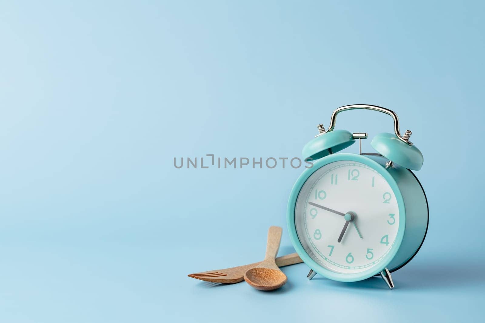 An alarm clock with cutlery set against blue background for the concept of food, time management, losing weight and eating on time.