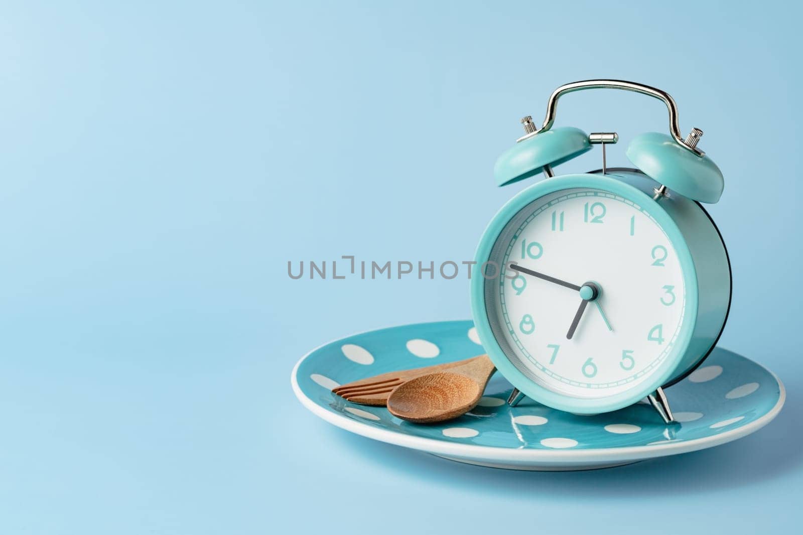 An alarm clock on an empty plate and cutlery set against blue background for the concept of food, time management, losing weight and eating on time.