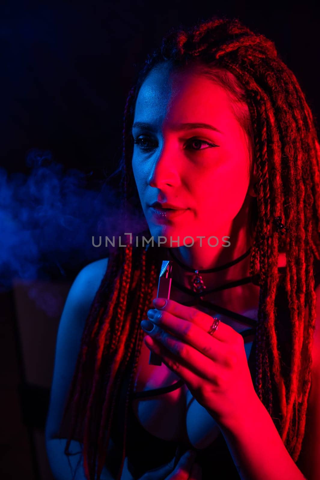 Caucasian girl with dreadlocks smokes a vape in red blue light