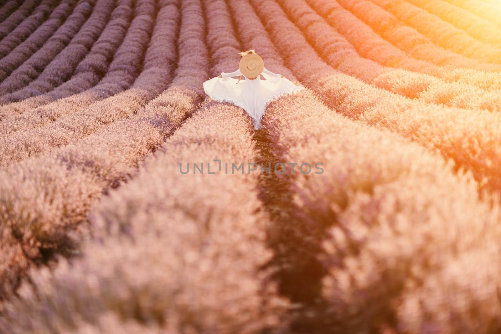 Happy woman in a white dress and straw hat strolling through a lavender field at sunrise, taking in the tranquil atmosphere