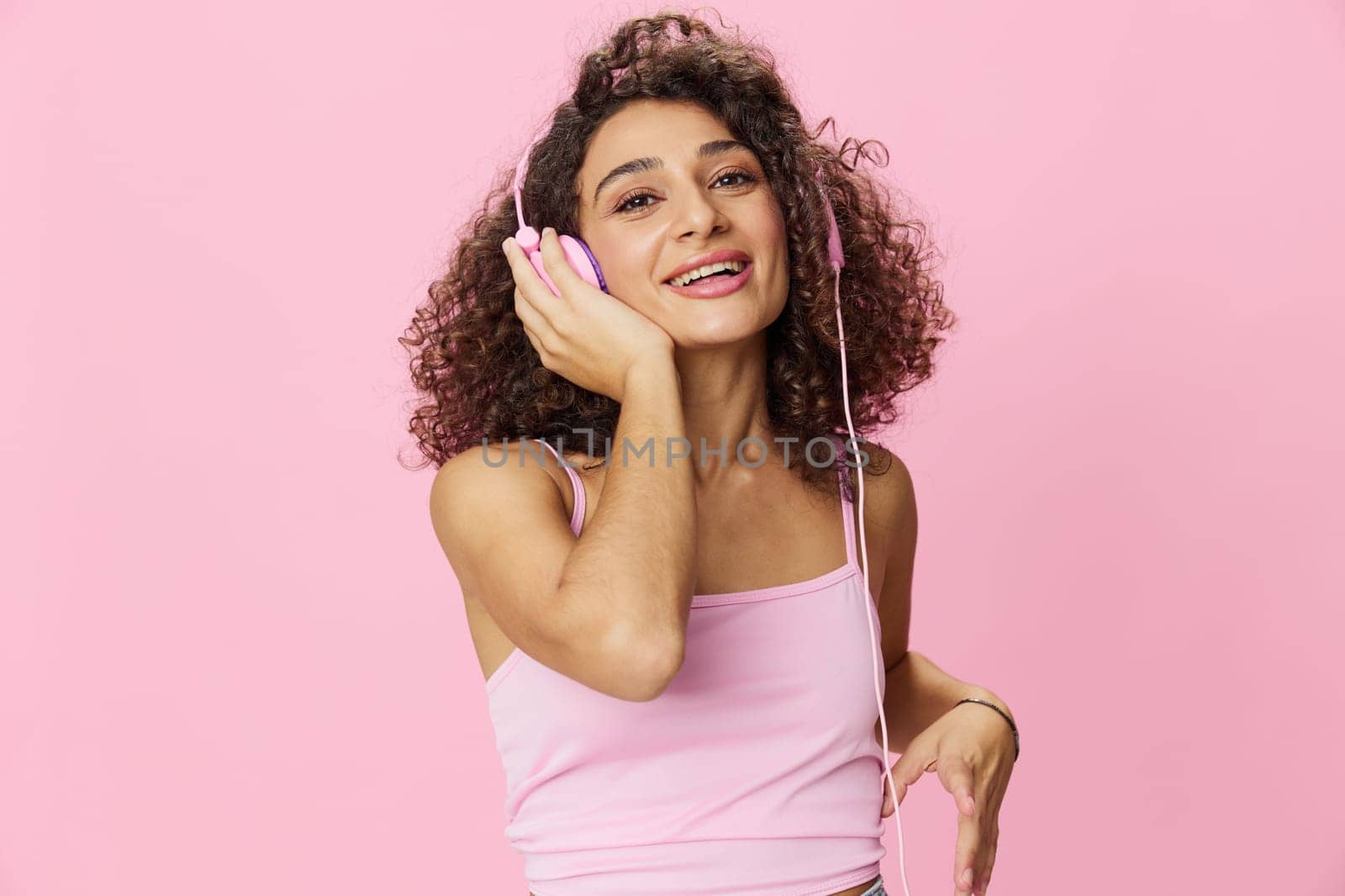Happy woman wearing headphones with curly hair listening to music and singing along with her eyes closed in a pink T-shirt and jeans on a pink background DJ party, copy space by SHOTPRIME