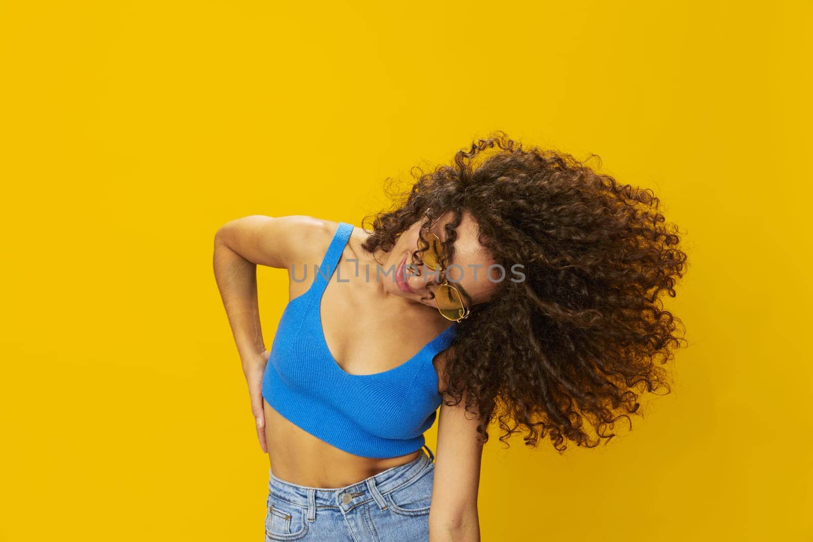 Woman with curly afro hair in a blue T-shirt on a yellow background dancing flying hair with sunglasses yellow, hand signs, look into the camera, smile with teeth and happiness, copy space. High quality photo