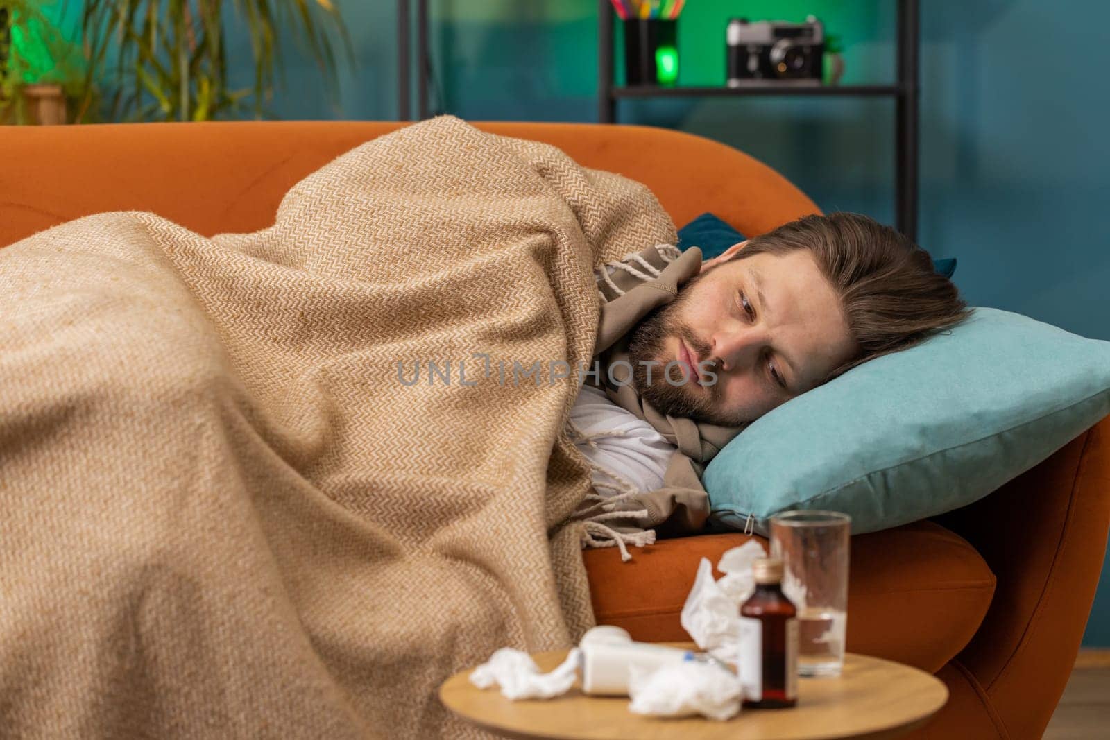 Young ill man suffering from cold or allergy lying on sofa in living room at home apartment. Sick Caucasian guy with towel on head blows sneezes wipes snot into napkin. Coronavirus quarantine pandemic