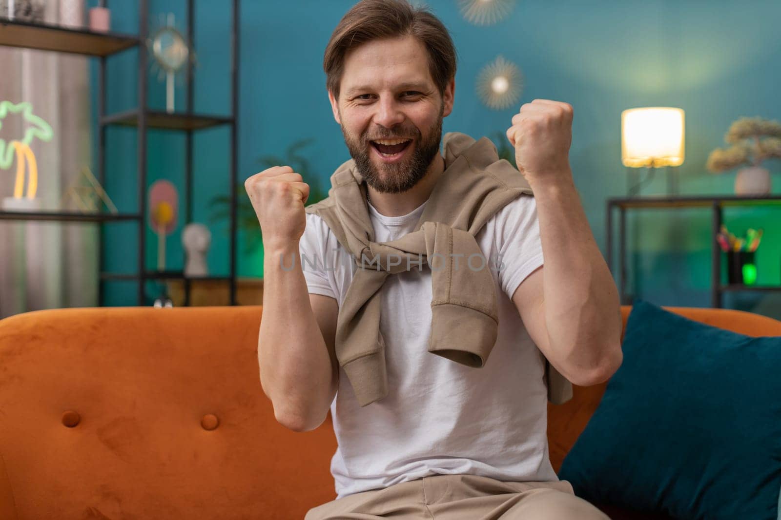 Happy Caucasian man shouting, celebrating success, winning, goal achievement good news, lottery luck, victory, watching TV championship at modern home apartment. Guy in living room sitting on couch