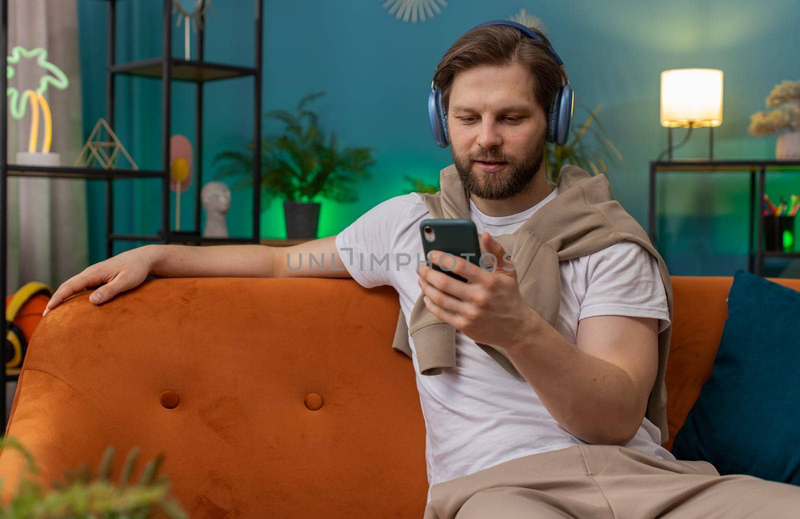Happy overjoyed Caucasian man in wireless headphones relaxing sitting on couch at home apartment choosing listening favorite energetic disco dancing music. People weekend daytime leisure activities