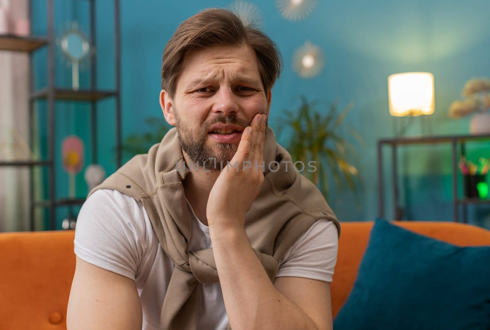 Dental problems. Young man touching cheek, closing eyes with expression of terrible suffer from painful toothache, sensitive teeth, cavities. Caucasian guy at home apartment living room on orange sofa