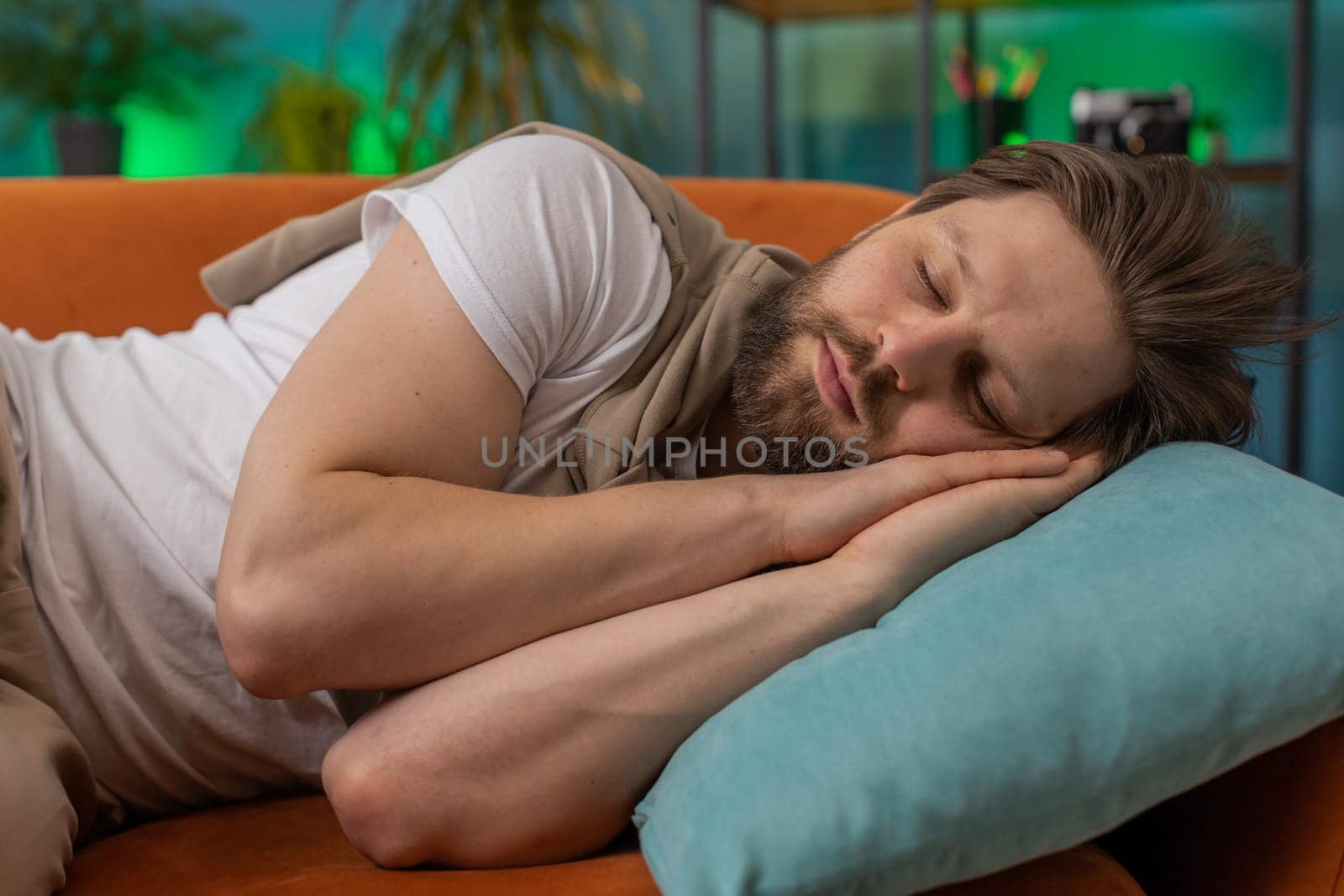 Tired young man lying down in bed taking a rest at home apartment room. Carefree Caucasian guy napping, falling asleep on comfortable orange sofa with pillows. Closed his eyes enjoy daytime nap alone