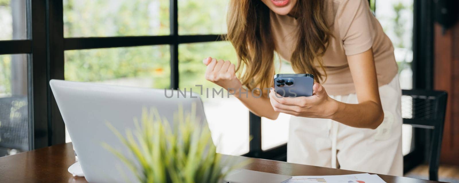 Excited woman holding mobile smart phone raising hands after playing online game and making winner gesture, young female smile got good news on smartphone while working on computer at cefa