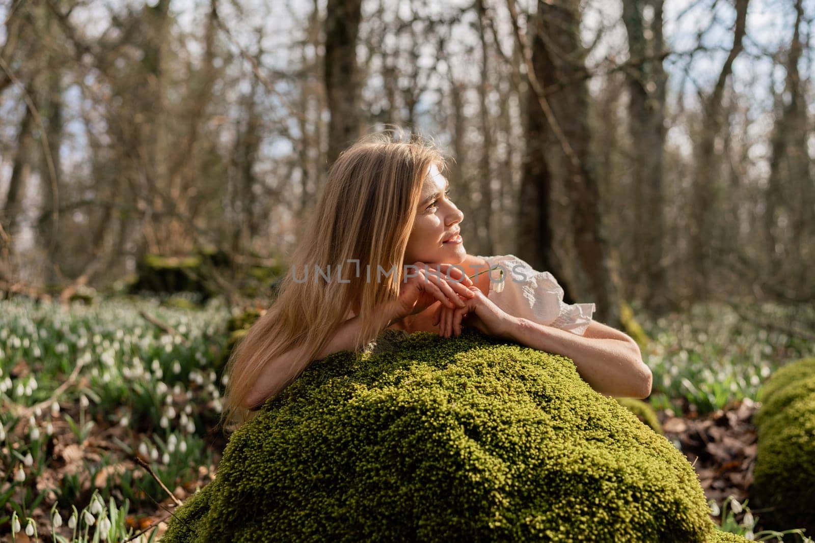 Snowdrops galanthus blond. A girl in a white dress lay down on a stone in the moss in a meadow with snowdrops in a spring forest by Matiunina