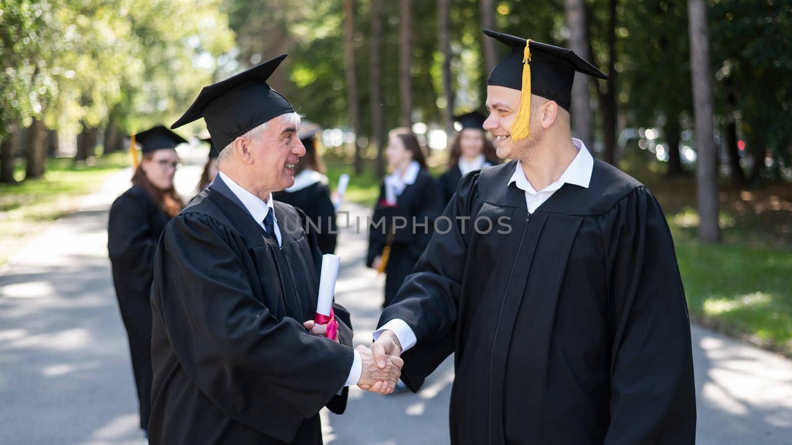A group of graduates in robes outdoors. An elderly man and a young guy congratulate each other on receiving a diploma
