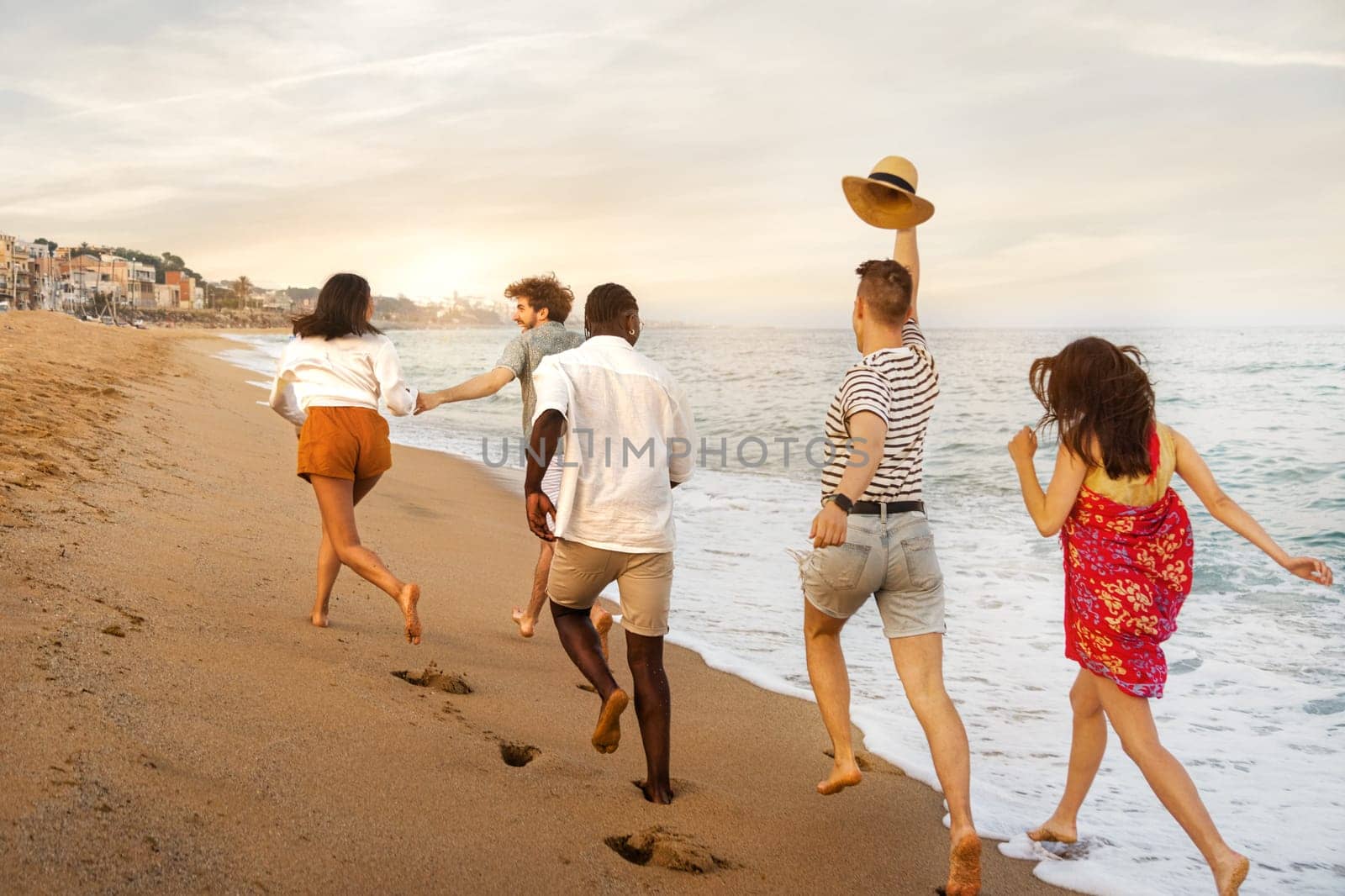 Rear view of group of young multiracial happy friends running along seaside during vacation travel. Holiday concept.
