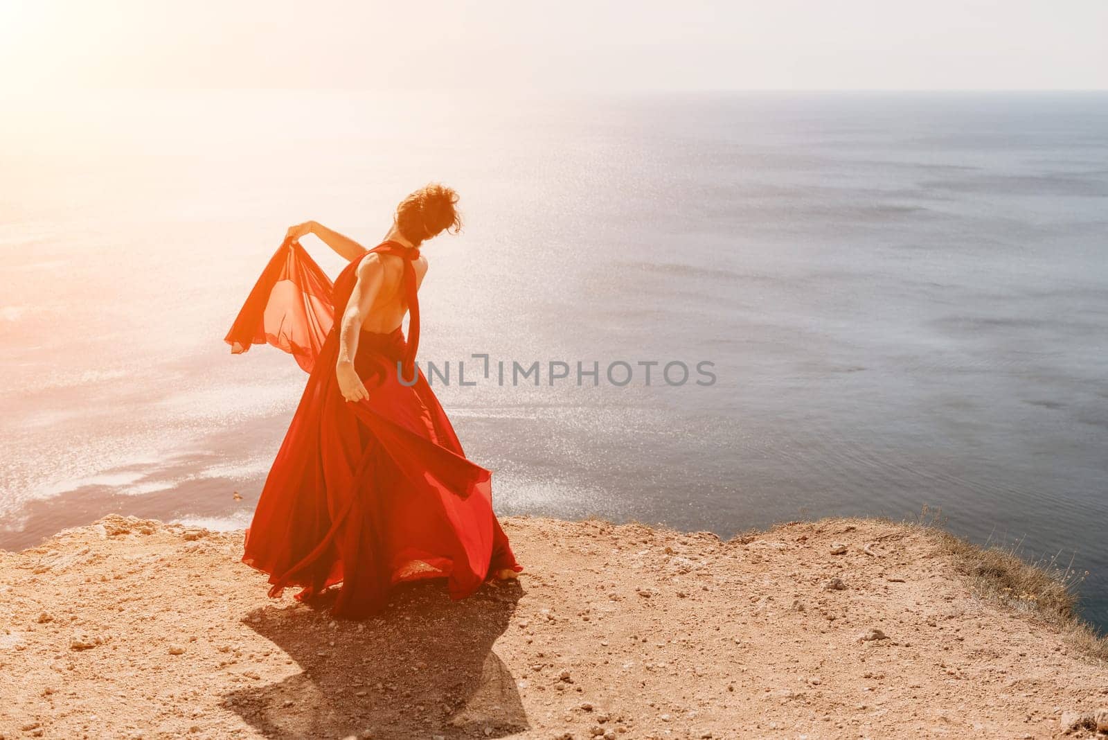 Side view a Young beautiful sensual woman in a red long dress posing on a rock high above the sea during sunrise. Girl on the nature on blue sky background. Fashion photo.