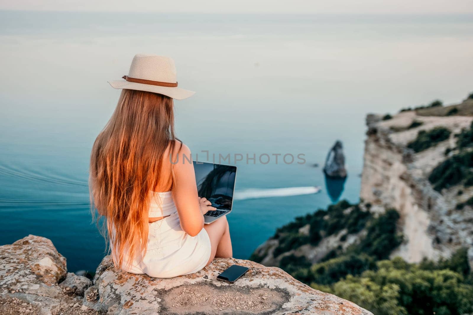 Woman laptop sea. Working remotely on seashore. Happy successful woman female freelancer in straw hat working on laptop by the sea at sunset. Freelance, remote work on vacation by panophotograph