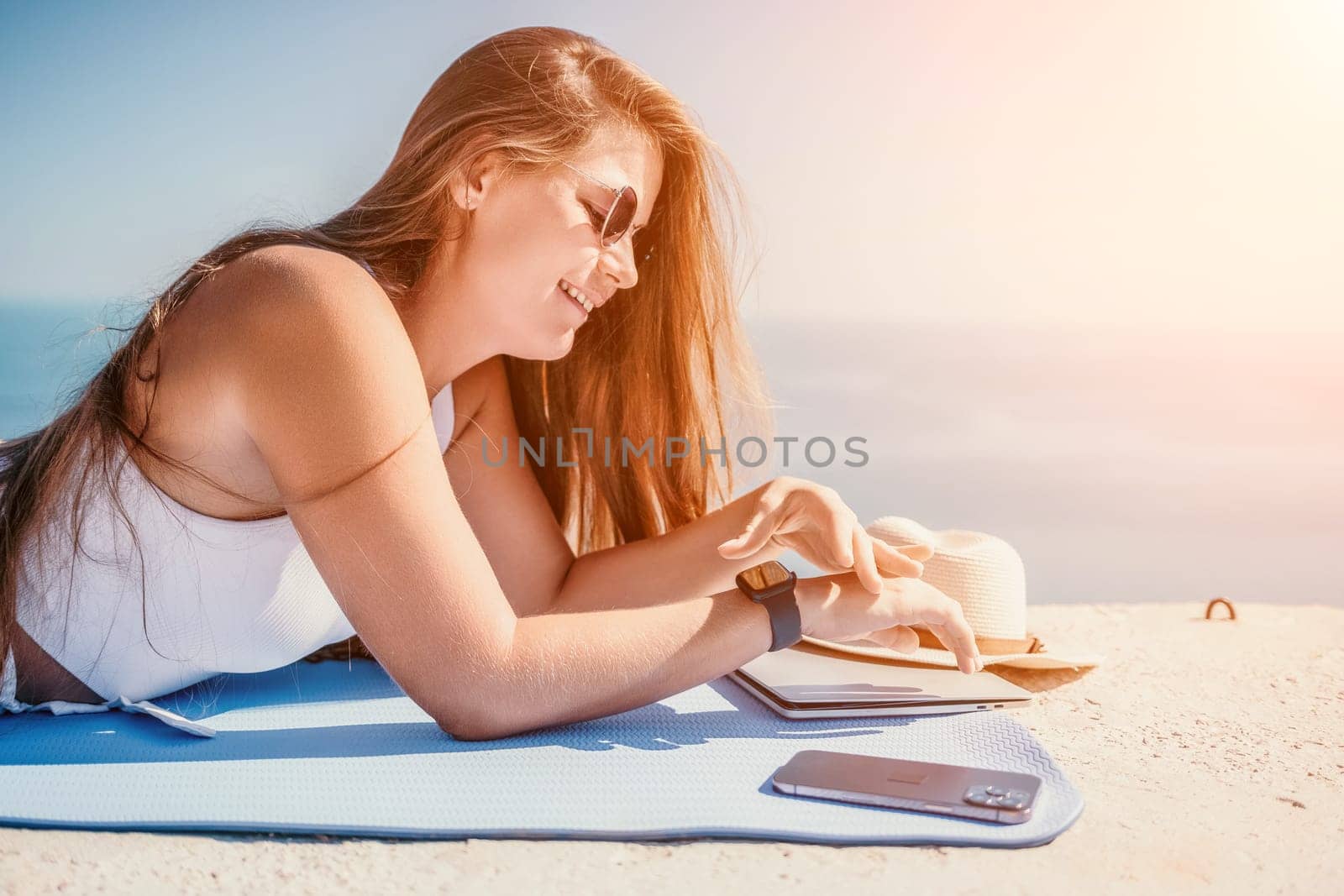 Digital nomad, woman in the hat, a business woman with a laptop sits on the rocks by the sea during sunset, makes a business transaction online from a distance. Freelance, remote work on vacation. by panophotograph