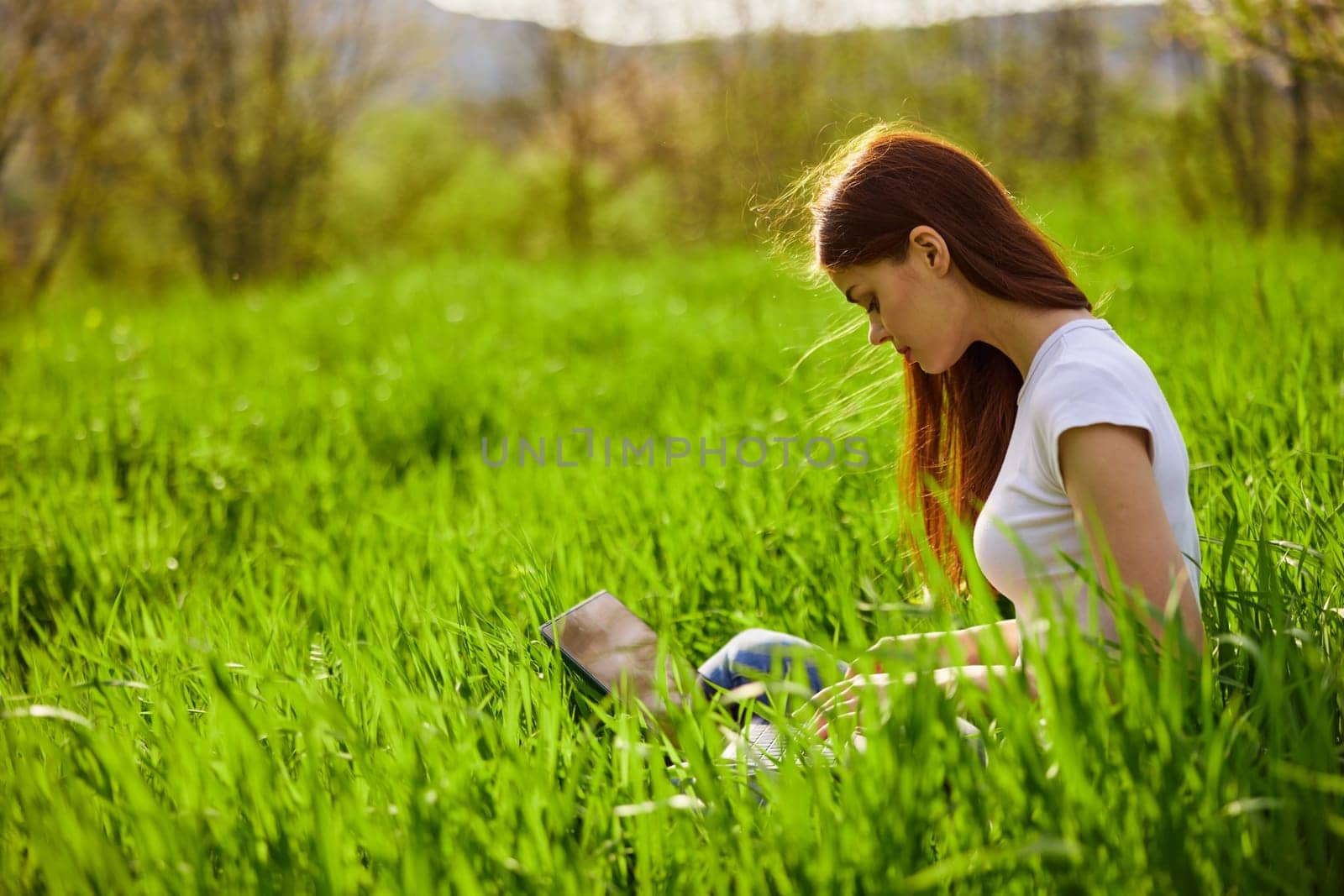 woman working on a laptop while sitting in the tall grass on the street by Vichizh