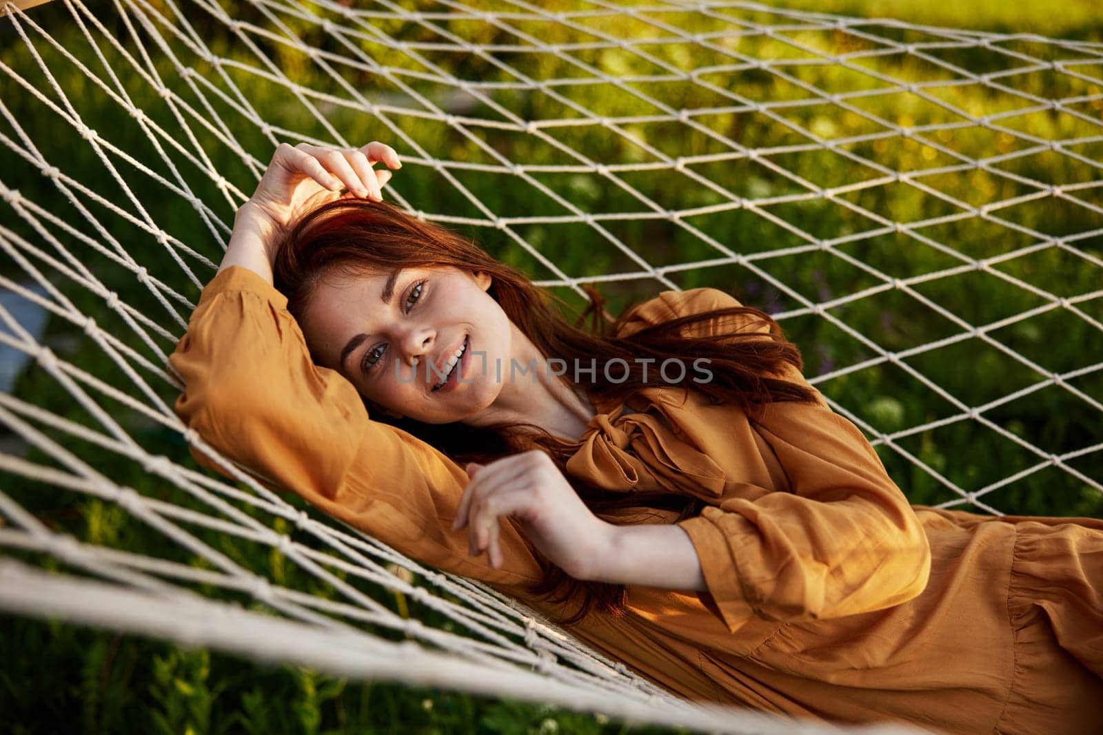 a happy woman is resting in a mesh hammock with her head resting on her hand, smiling happily at the camera with a smile, enjoying a warm day in the rays of the setting sun, lying in an orange dress. High quality photo