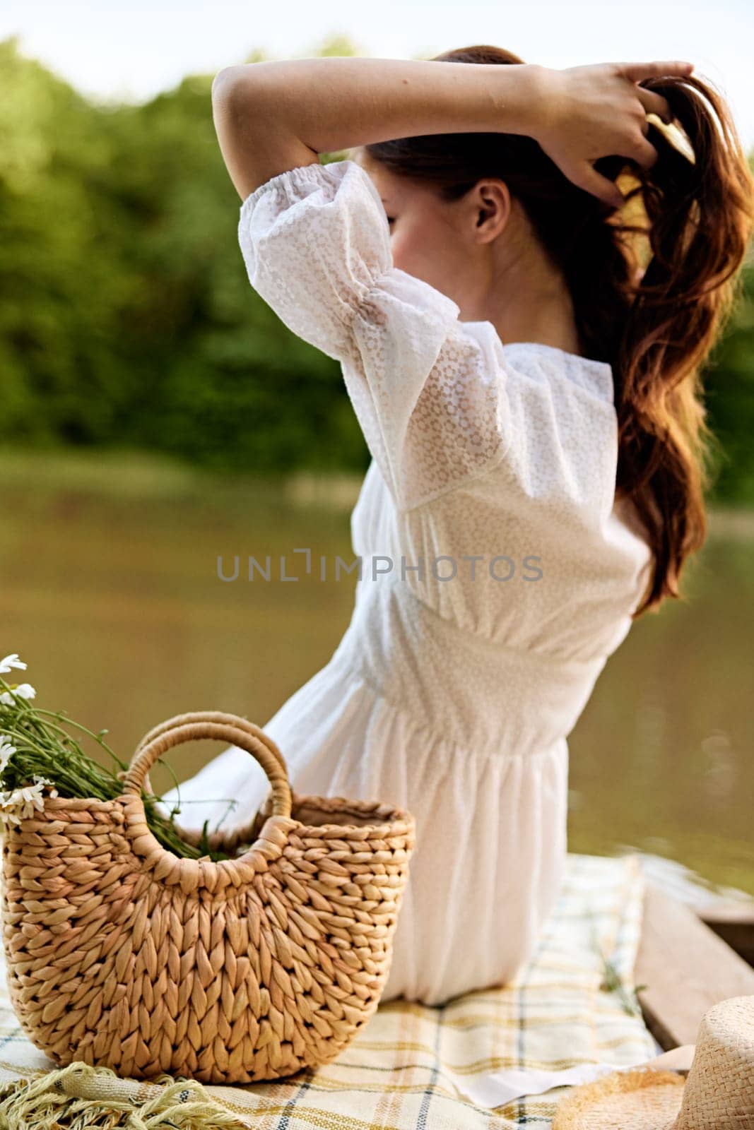 close-up photo of a woman sitting with her back to the camera on the shore of a lake with a wicker bag full of daisies. High quality photo