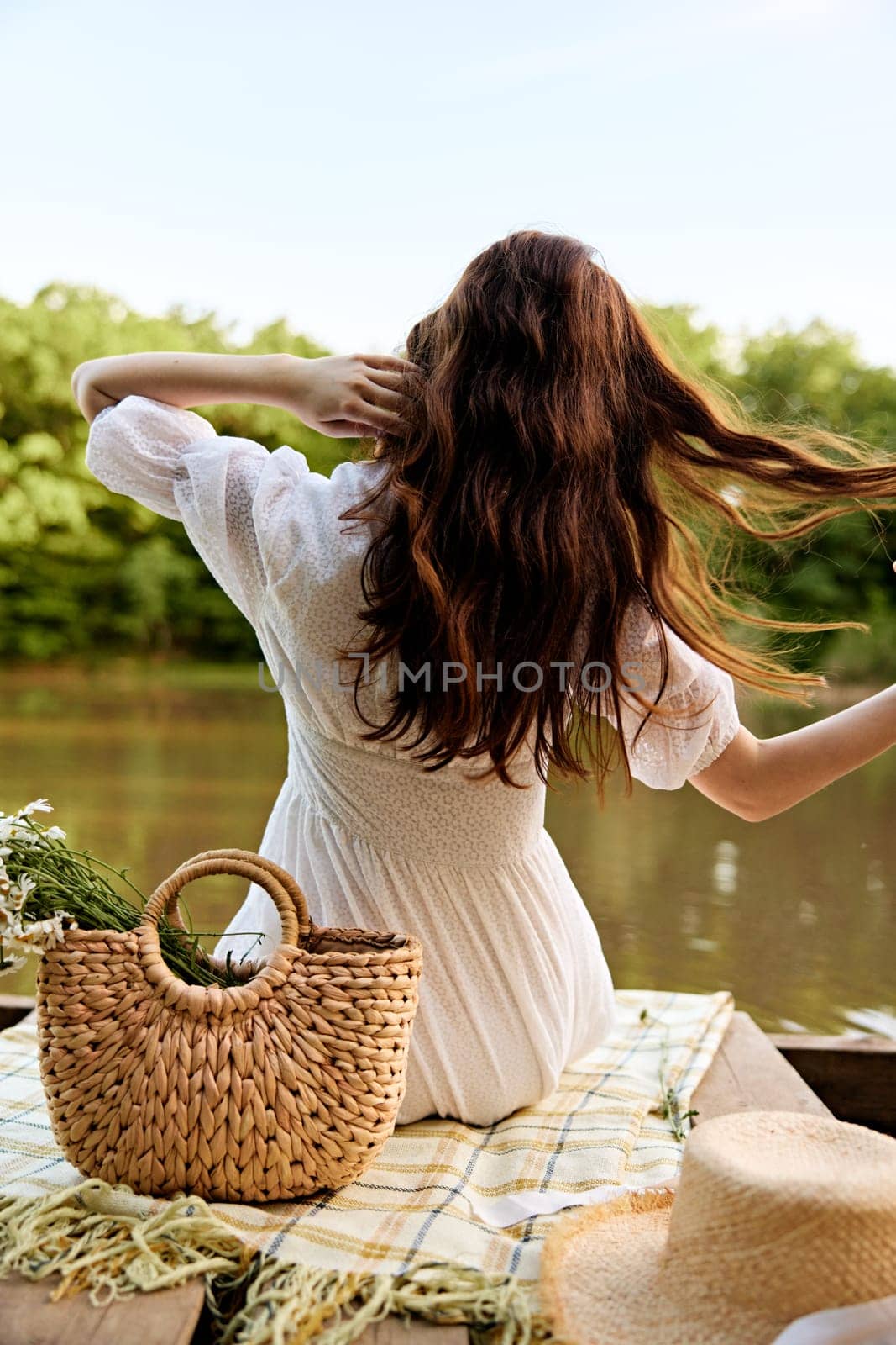 close-up photo of a woman sitting with her back to the camera on the shore of a lake with a wicker bag full of daisies. High quality photo