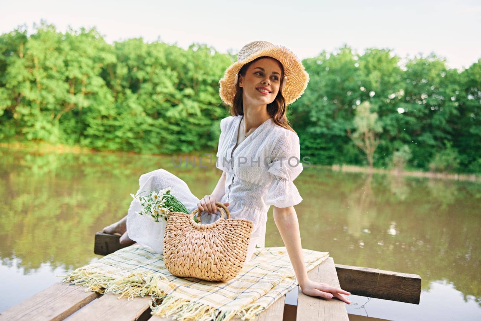 a beautiful woman in a light, summer dress and a wicker hat is resting on a pier by the lake, sitting on a plaid by Vichizh