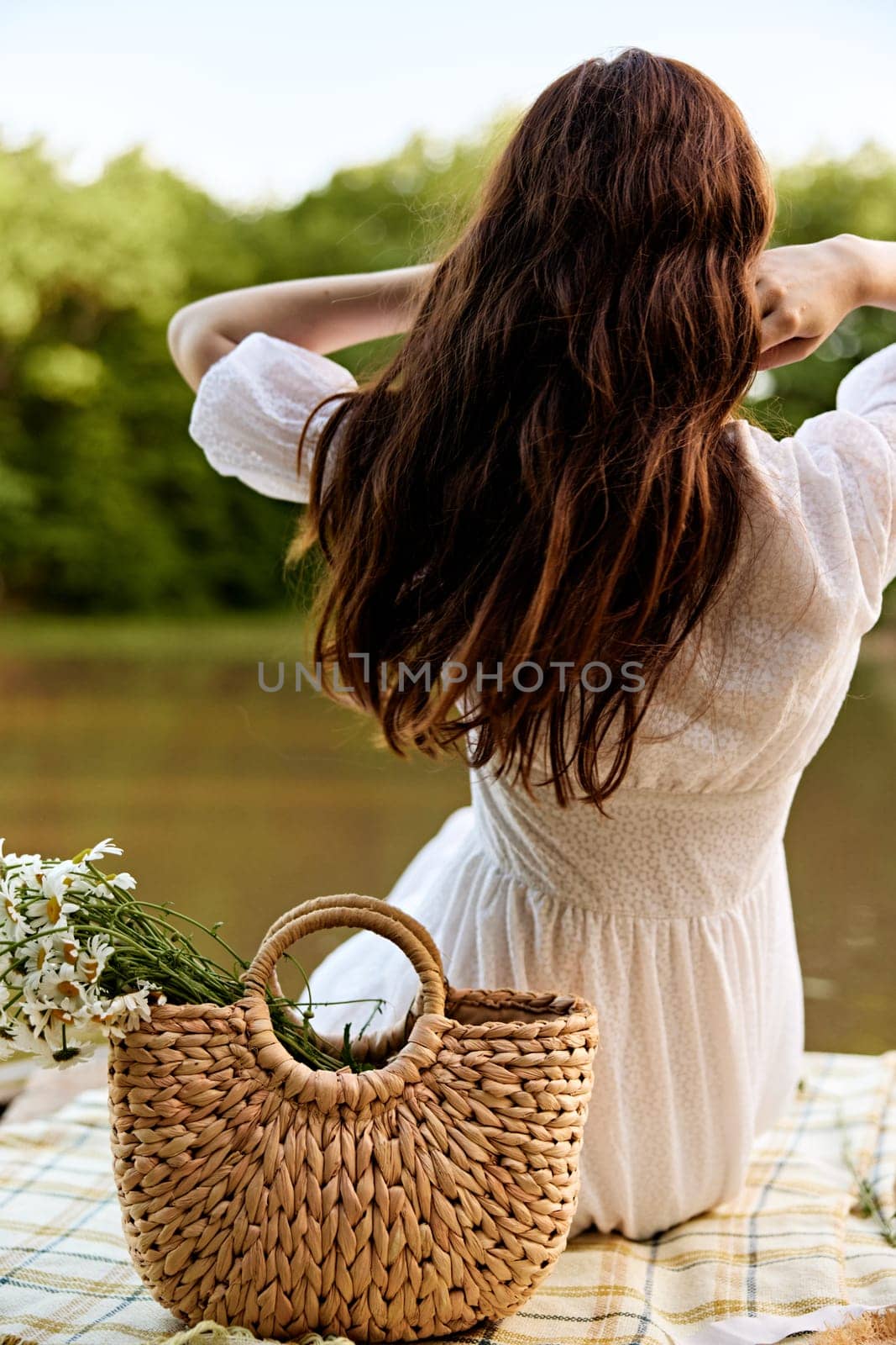close-up photo of a woman sitting with her back to the camera on the shore of a lake with a wicker bag full of daisies. High quality photo