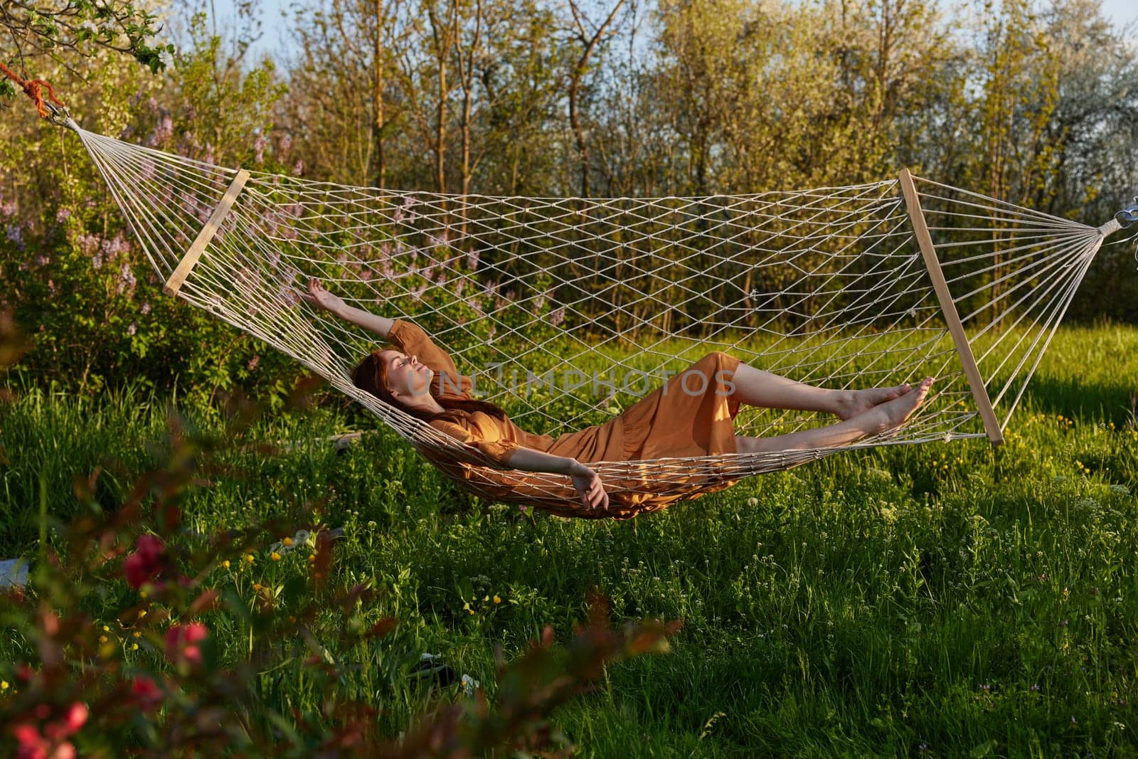 a happy woman in a long orange dress is relaxing in nature lying in a mesh hammock enjoying summer and vacation, looking at the sky by Vichizh