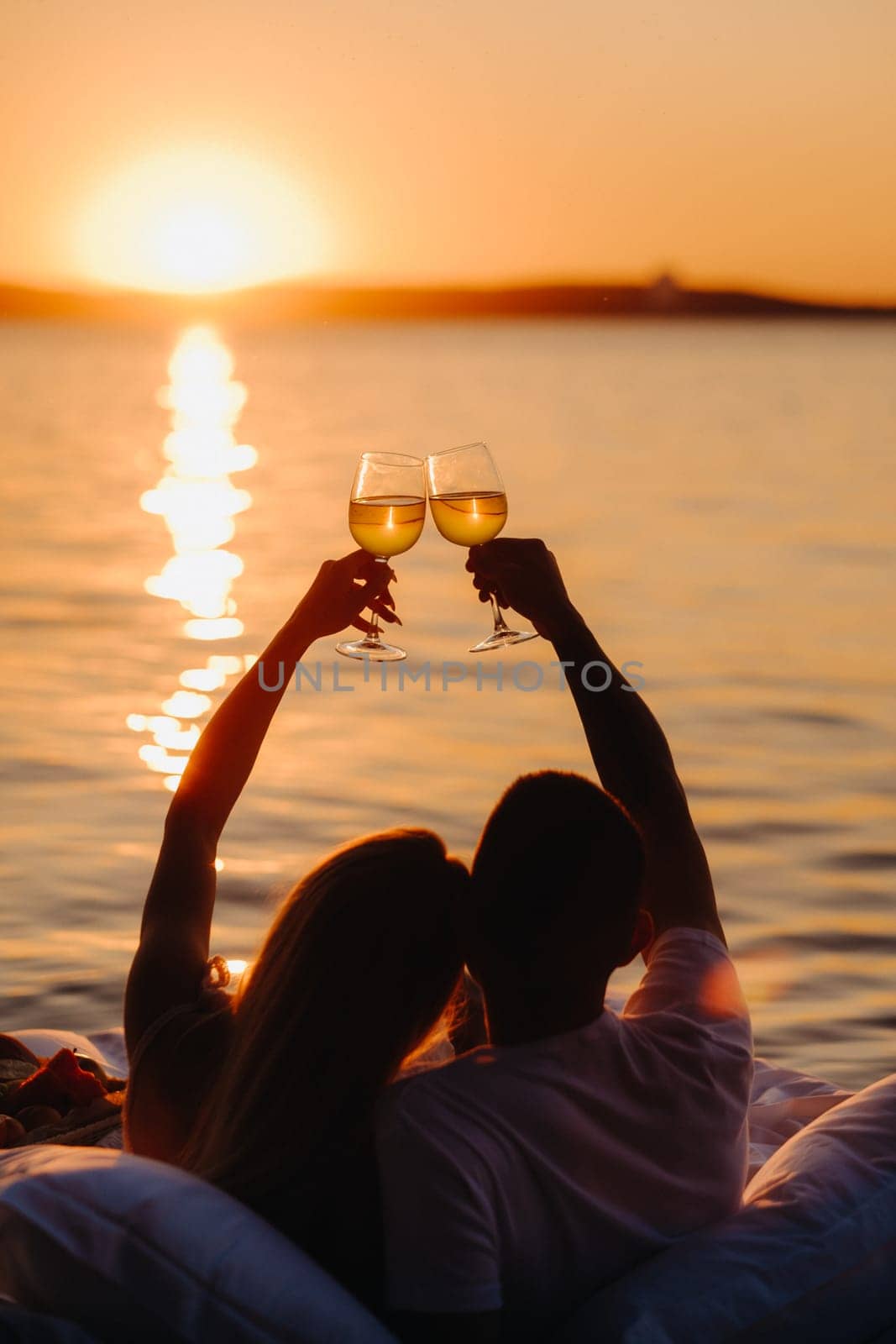 Silhouettes of a happy couple raising glasses on a summer evening near the sea at sunset.