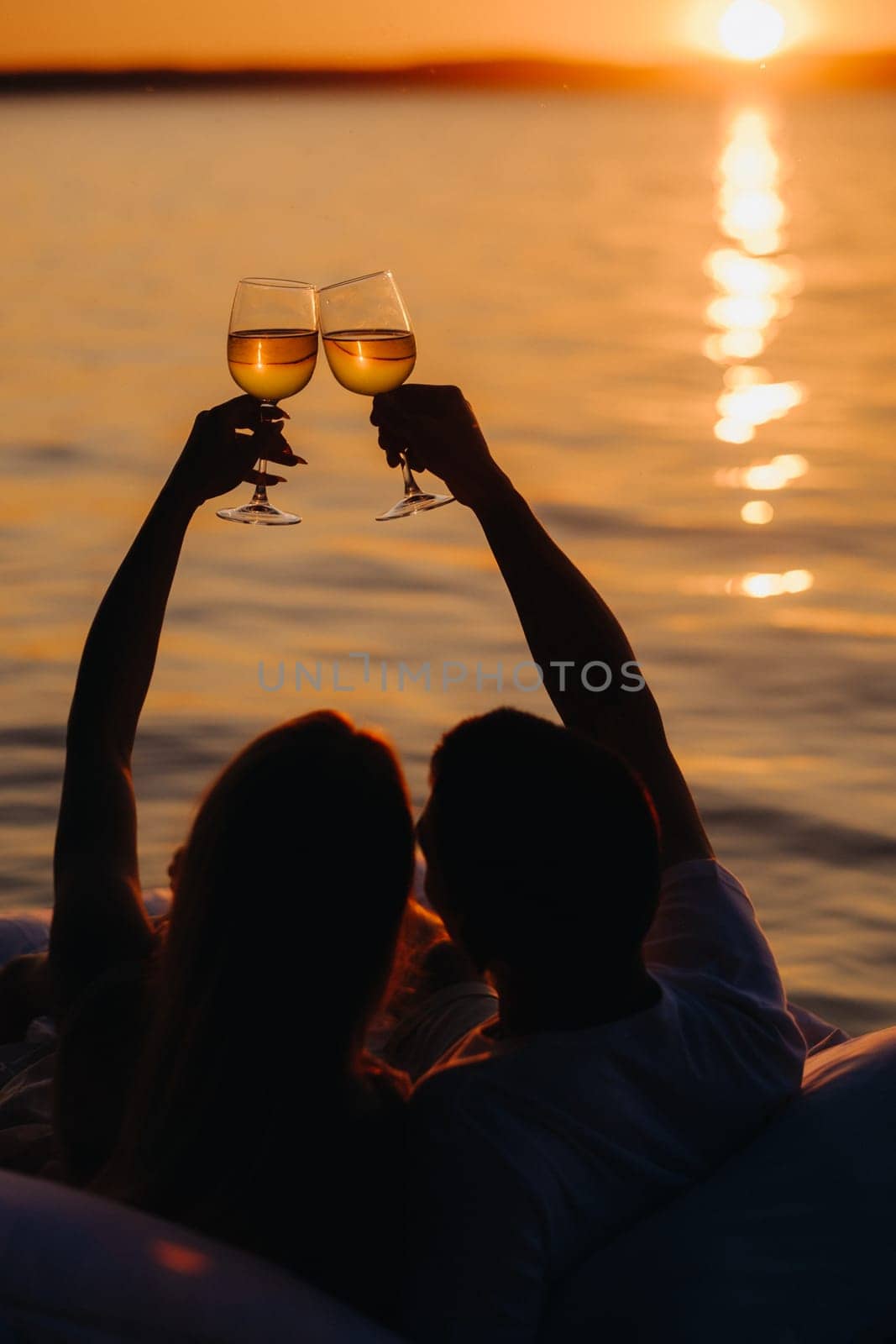 Silhouettes of a happy couple raising glasses on a summer evening near the sea at sunset.