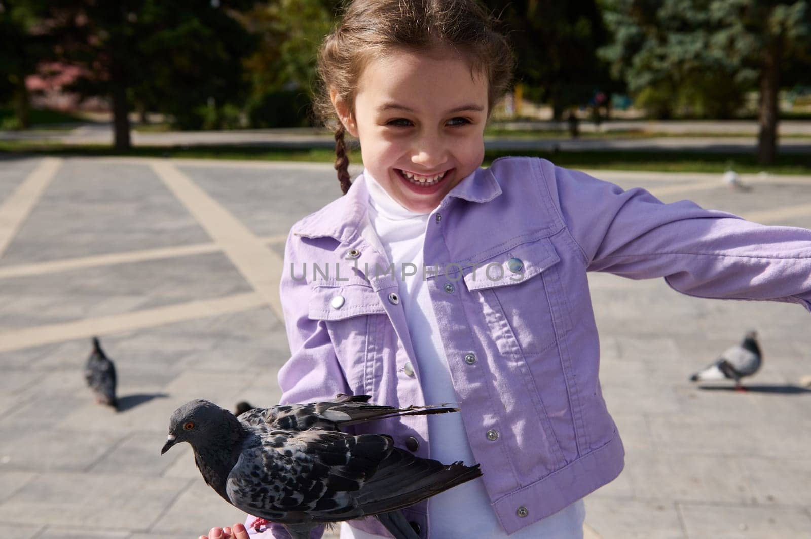 Happy overjoyed Caucasian little child girl rejoices at a pigeon sitting on her arm, smiling, expressing happiness while feeding flock of flying birds in the city park on a sunny spring day