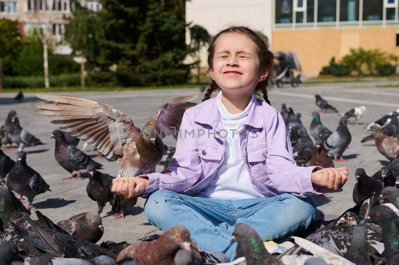 Happy Caucasian adorable lovely little child girl, sitting in lotus pose, meditating with her eyes closed while feeding flock of flying pigeons and birds in the park square, on a warm sunny spring day