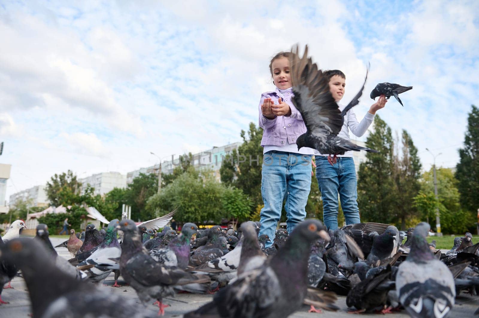 Adorable children boy and girl, feeding rock pigeons crowding the street with discarded food in the city square. The concept of happy carefree childhood and instilling love for nature and animals