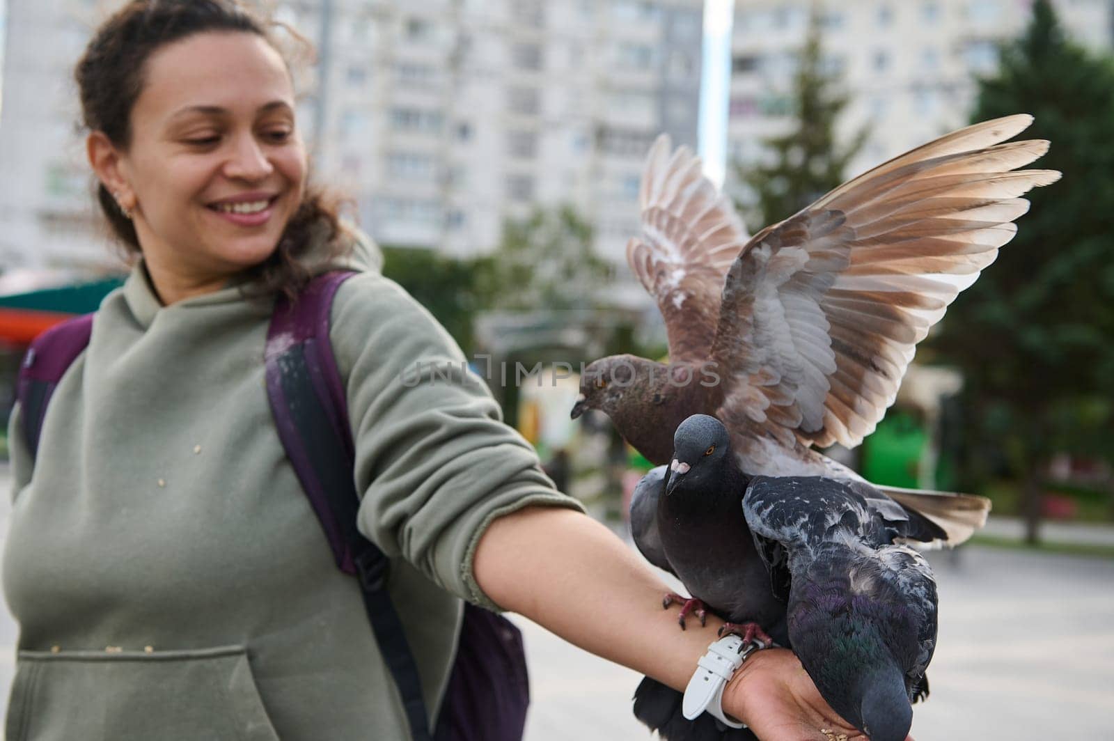 Selective focus on feral doves, rock pigeons on outstretched arm of a smiling positive woman feeding birds in the park by artgf