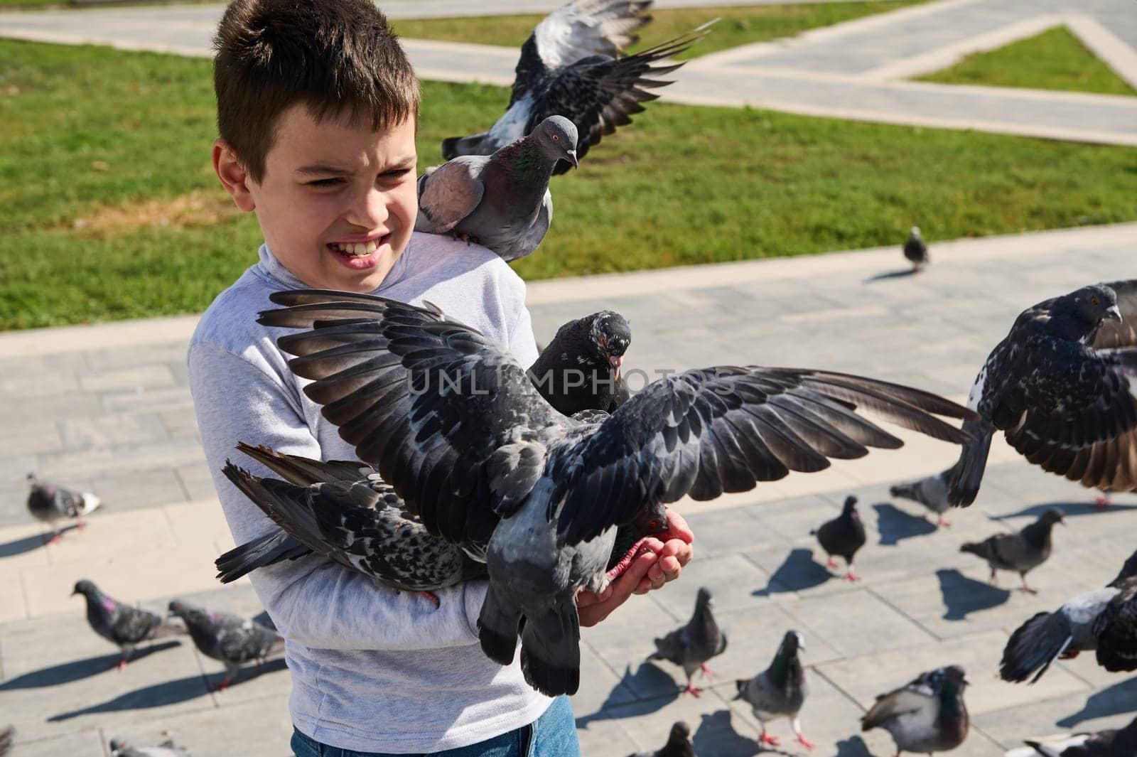 Caucasian teen boy feeding flock of flying pigeons in the summer city park square. Children and wild animals by artgf