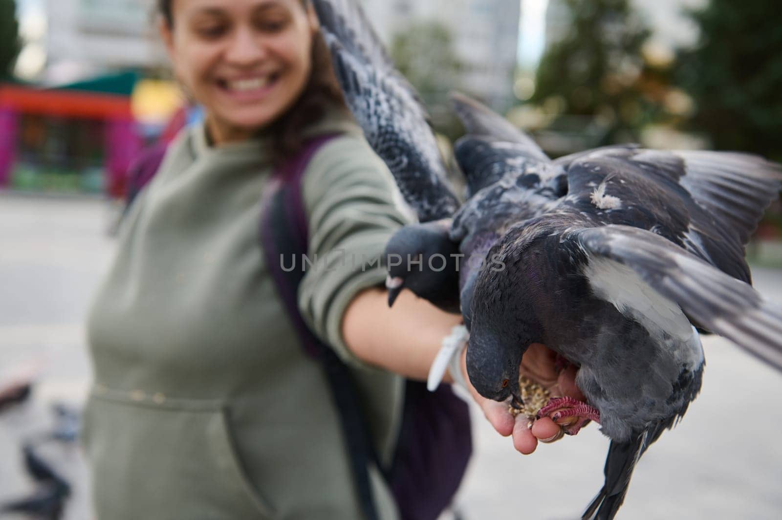 Selective focus on rock pigeons, sitting on the outstretched hand of a smiling African American positive woman feeding doves while outing in the square of a city park. Care and kindness for animals
