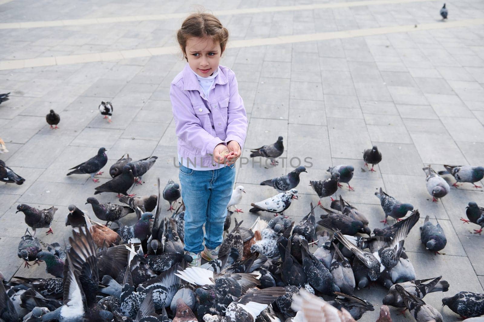 Full length portrait of a little girl feeding rock pigeons crowding the street and eating discarded food and bird seeds by artgf