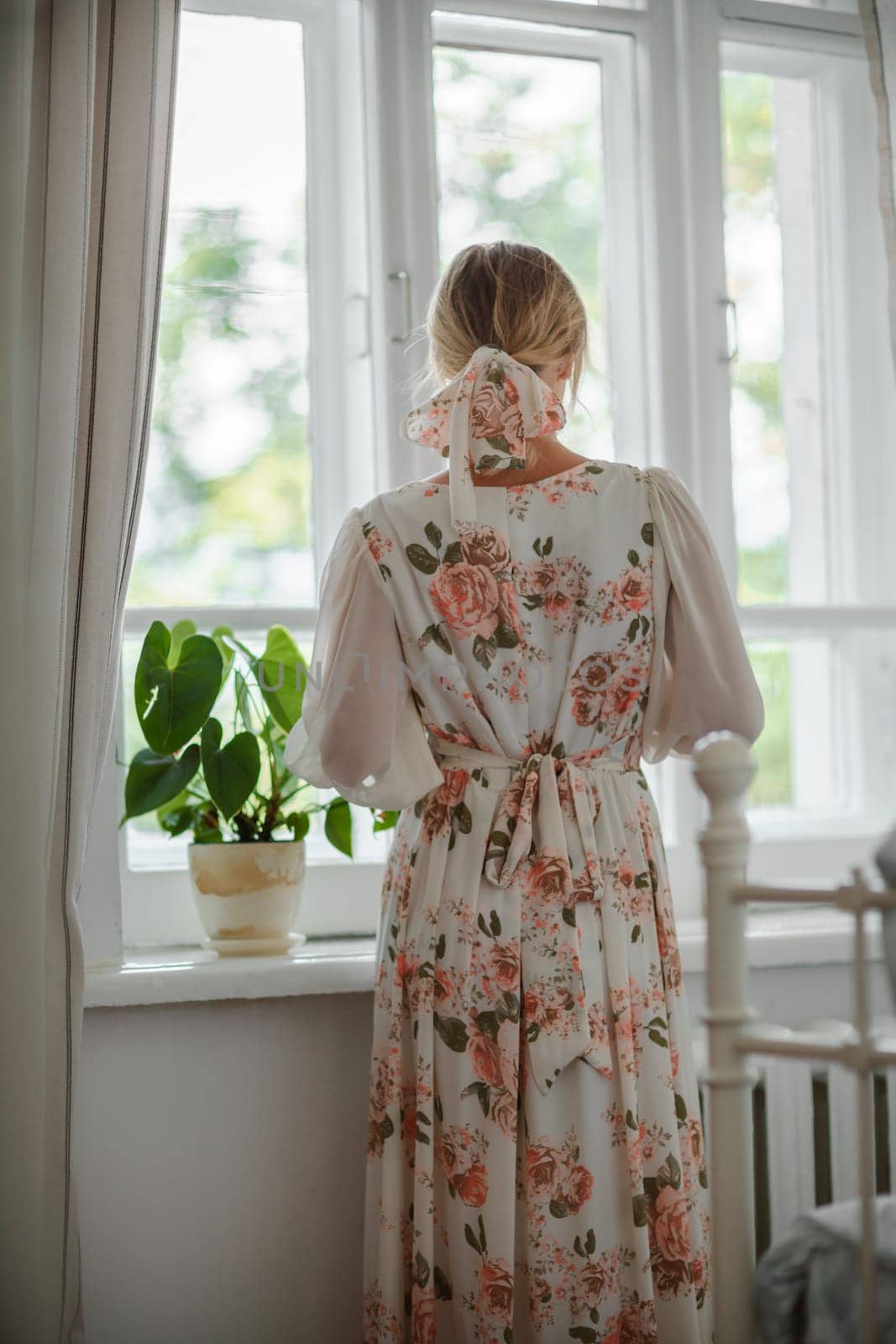 A woman wearing a cream dress with flowers stands by a window, lost in thought as she gazes at the scenery outside by Matiunina