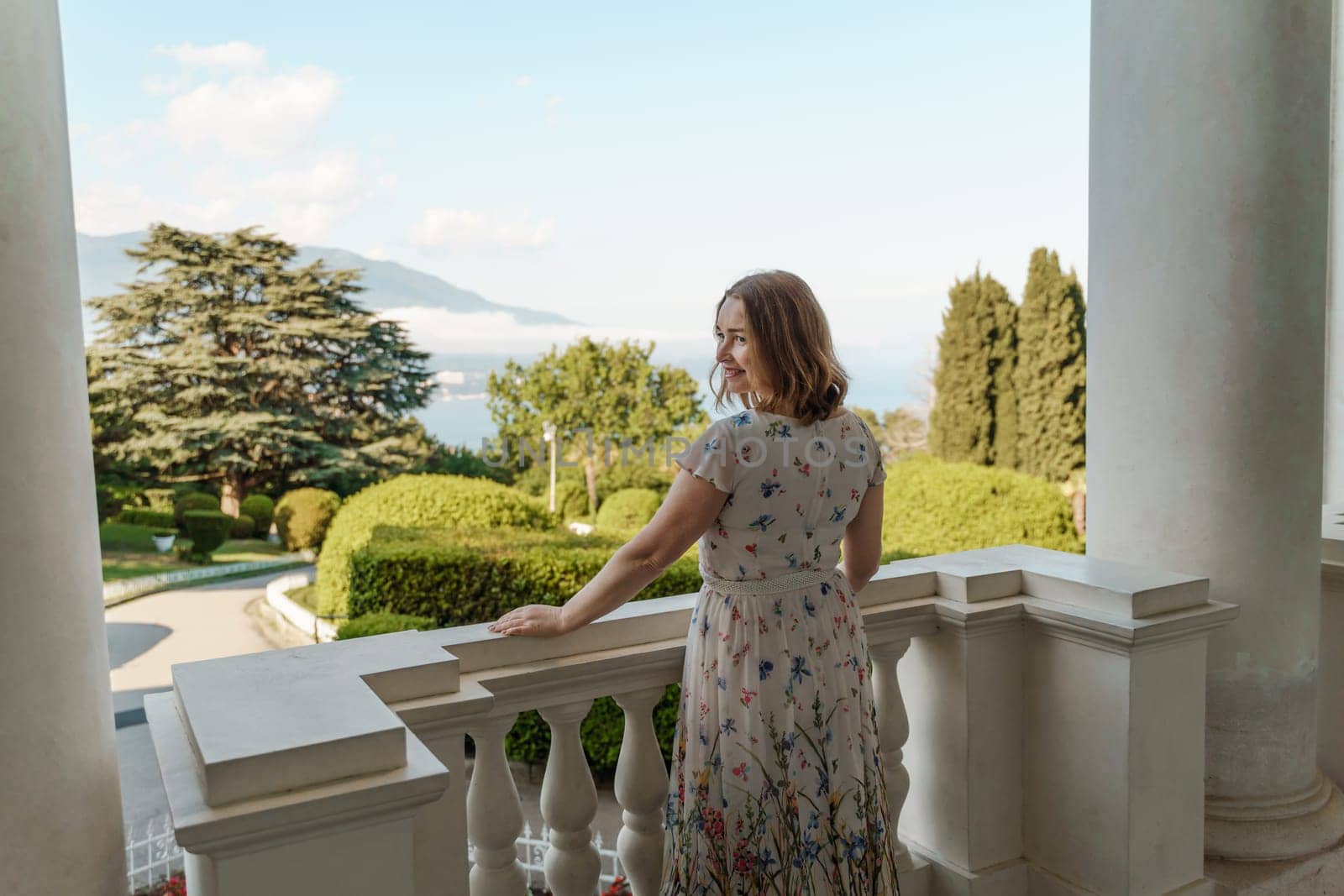 happy mature woman in a long white dress stands on the balcony and looks into the distance.