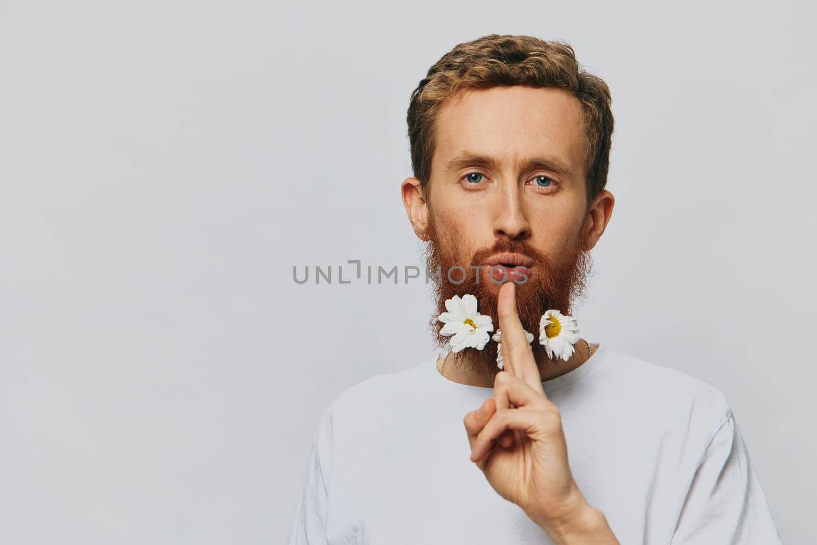 Portrait of a funny man in a white T-shirt with flowers daisies in his beard on a white isolated background, copy place. Holiday concept and congratulations. High quality photo