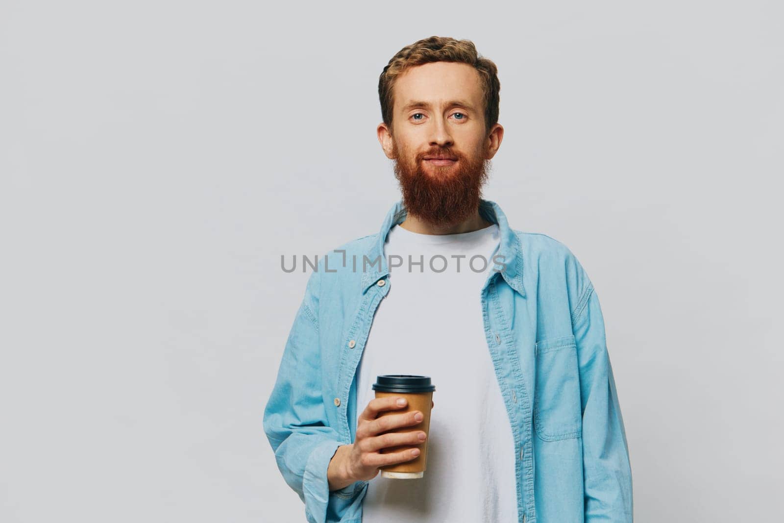 Man hipster with a cup of coffee with a smile on a gray background in a blue shirt and white t-shirt by SHOTPRIME