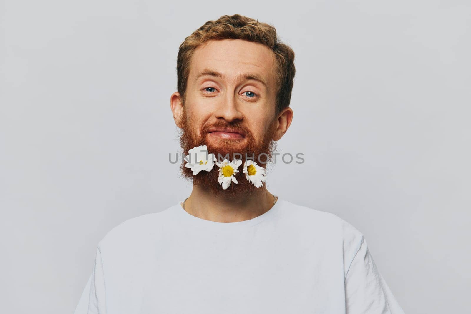 Portrait of a funny man in a white T-shirt with flowers daisies in his beard on a white isolated background, copy place. Holiday concept and congratulations. High quality photo