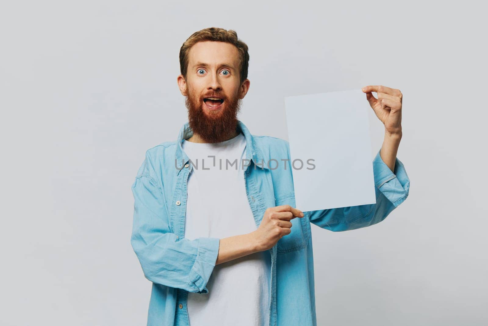 Portrait of an adult male on a gray background with a white sheet of paper in his hands for your design and text, layout, copy space, space for text, finger pointing by SHOTPRIME