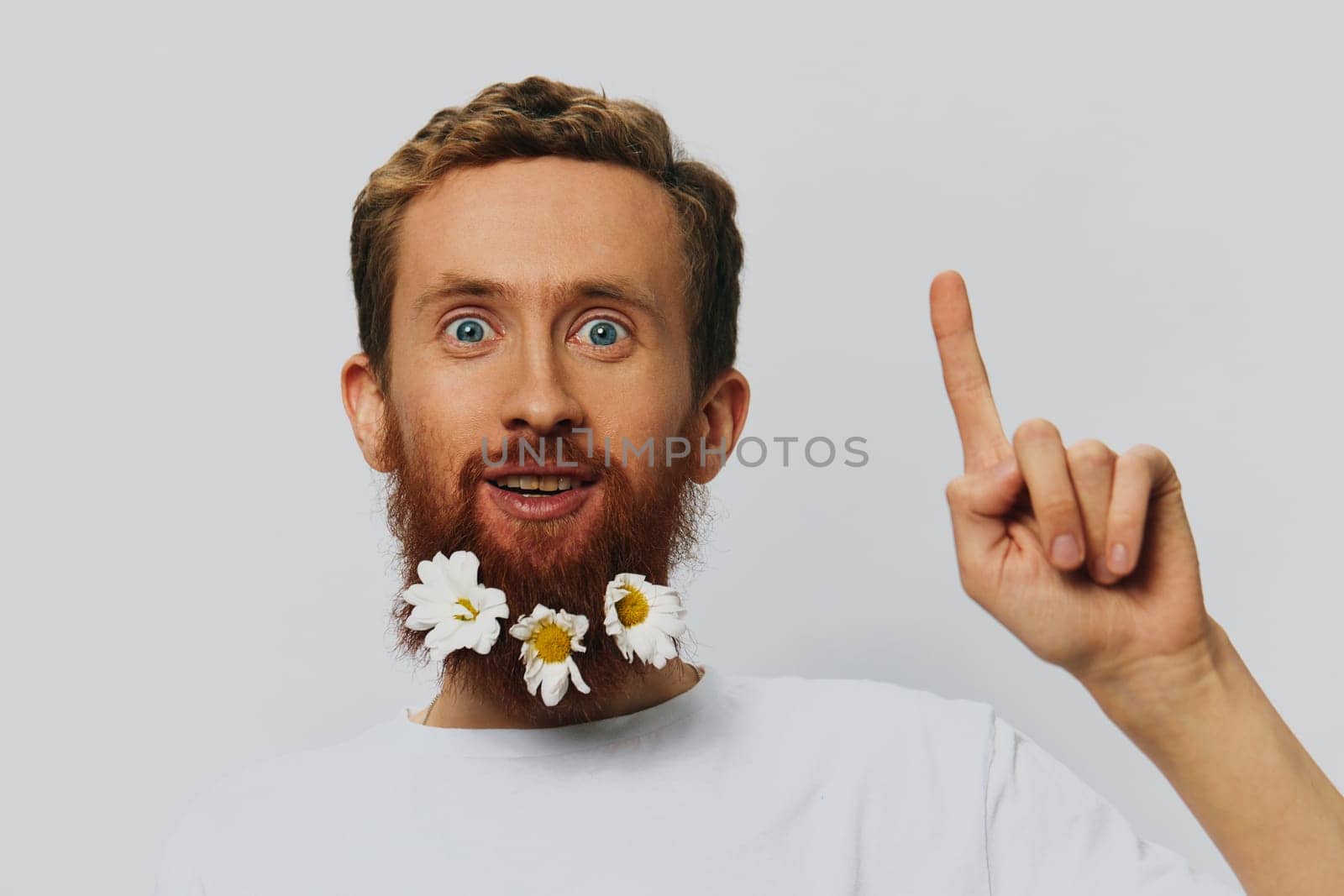 Portrait of a funny man in a white T-shirt with flowers daisies in his beard on a white isolated background, copy place. Holiday concept and congratulations. by SHOTPRIME