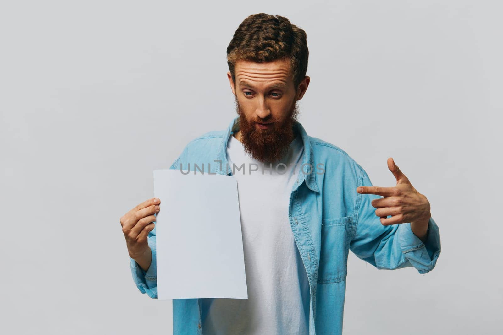 Portrait of an adult male on a gray background with a white sheet of paper in his hands for your design and text, layout, copy space, space for text, finger pointing by SHOTPRIME