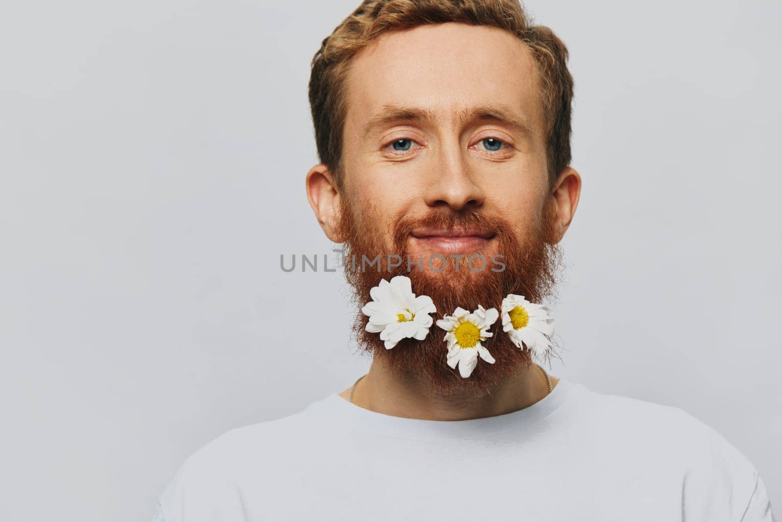 Portrait of a funny man in a white T-shirt with flowers daisies in his beard on a white isolated background, copy place. Holiday concept and congratulations. High quality photo