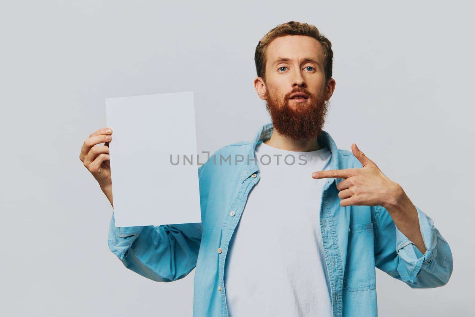 Portrait of an adult male on a gray background with a white sheet of paper in his hands for your design and text, layout, copy space, space for text, finger pointing by SHOTPRIME