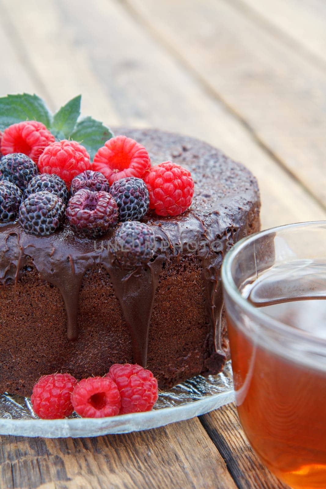 Homemade chocolate cake decorated with mint leaves, black and red raspberries on glass plate with cup of tea on wooden boards.