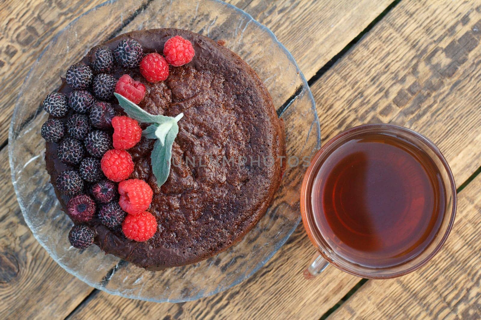 Homemade chocolate cake decorated with mint leaves, black and red raspberries on glass plate with cup of tea on wooden boards. Top view.