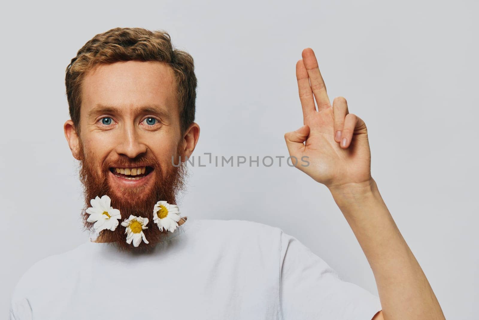 Portrait of a funny man in a white T-shirt with flowers daisies in his beard on a white isolated background, copy place. Holiday concept and congratulations. by SHOTPRIME