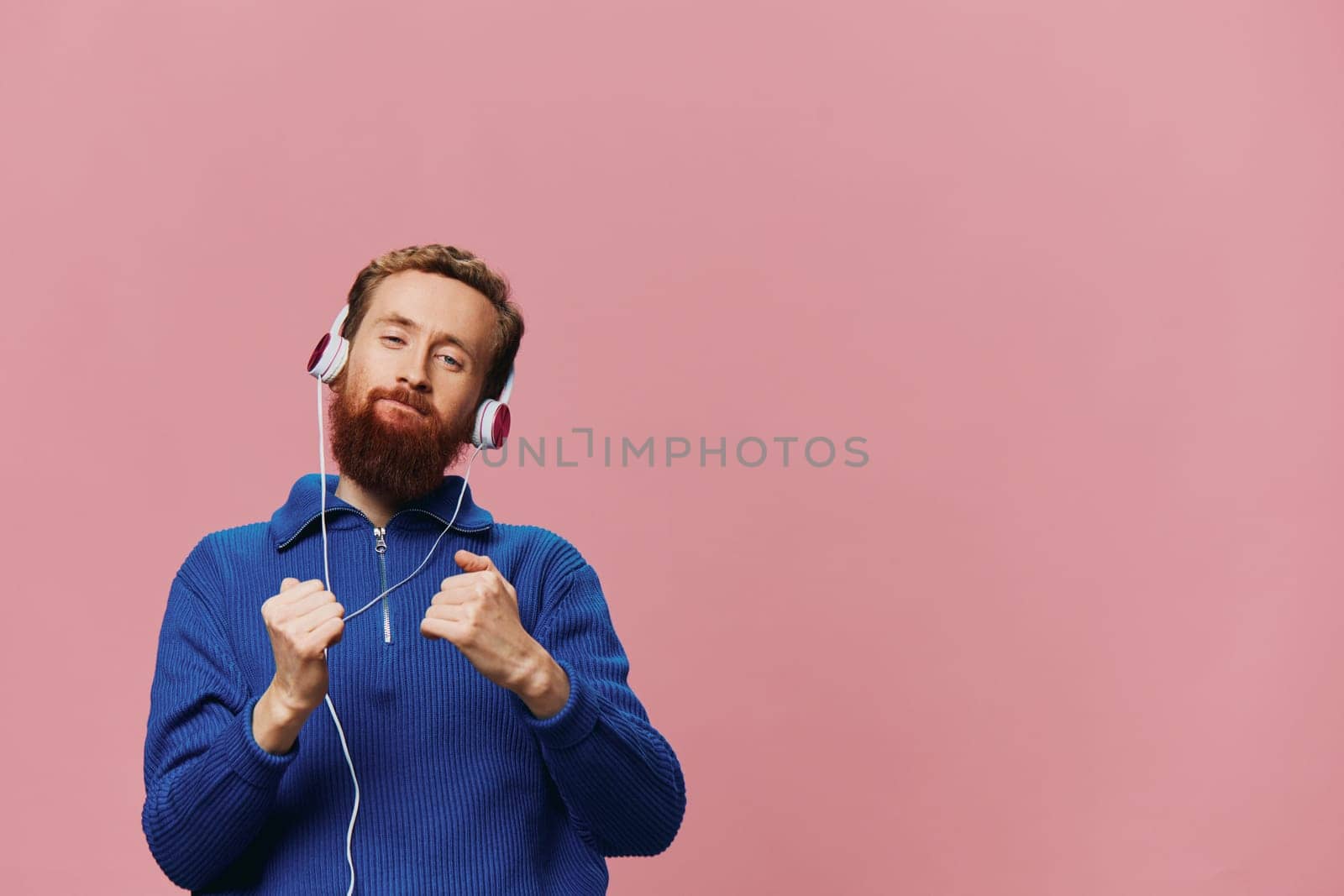 Portrait of a redheaded man wearing headphones smiling and dancing, listening to music on a pink background. A hipster with a beard. High quality photo