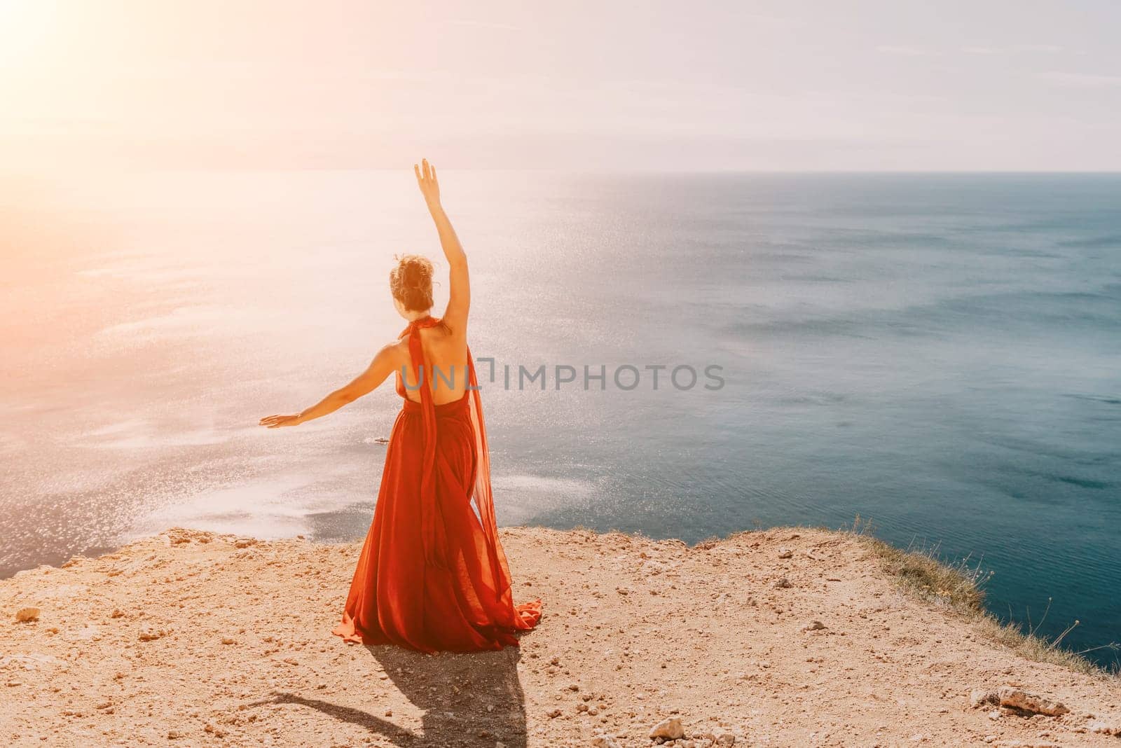 Side view a Young beautiful sensual woman in a red long dress posing on a rock high above the sea during sunrise. Girl on the nature on blue sky background. Fashion photo.